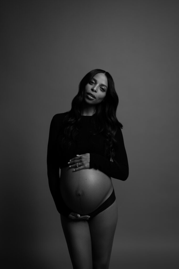 Studio maternity portrait of a Black pregnant woman in a black top and underwear, standing against a dark backdrop with a calm, direct gaze.