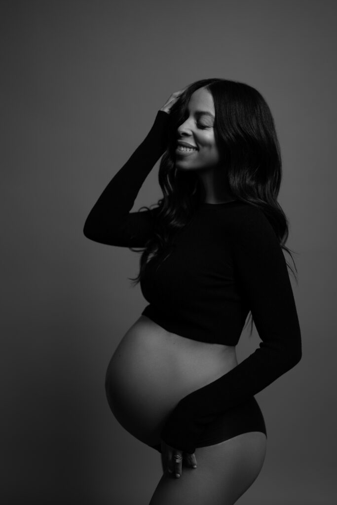 Black pregnant woman smiling joyfully with her eyes closed, hand in her hair, wearing a ribbed crop top and underwear in a studio setting.