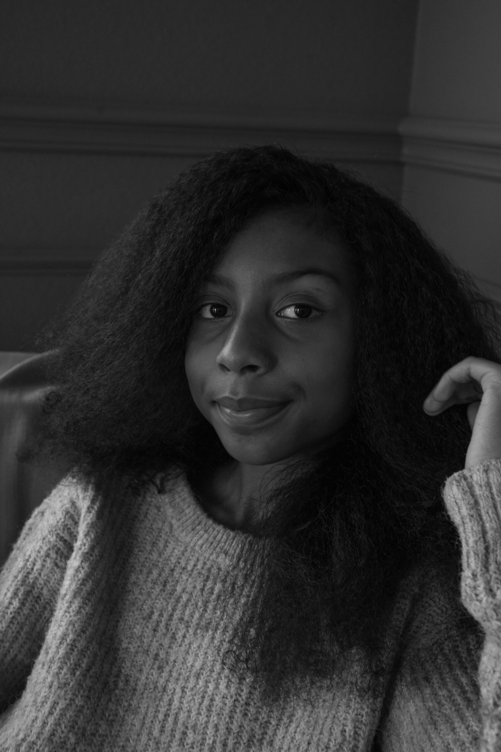 Black-and-white portrait of an 11-year-old girl seated against a textured wall, wearing a cozy knit sweater. Her calm expression and gentle smile convey a sense of quiet confidence and introspection, capturing the essence of her growth and individuality.