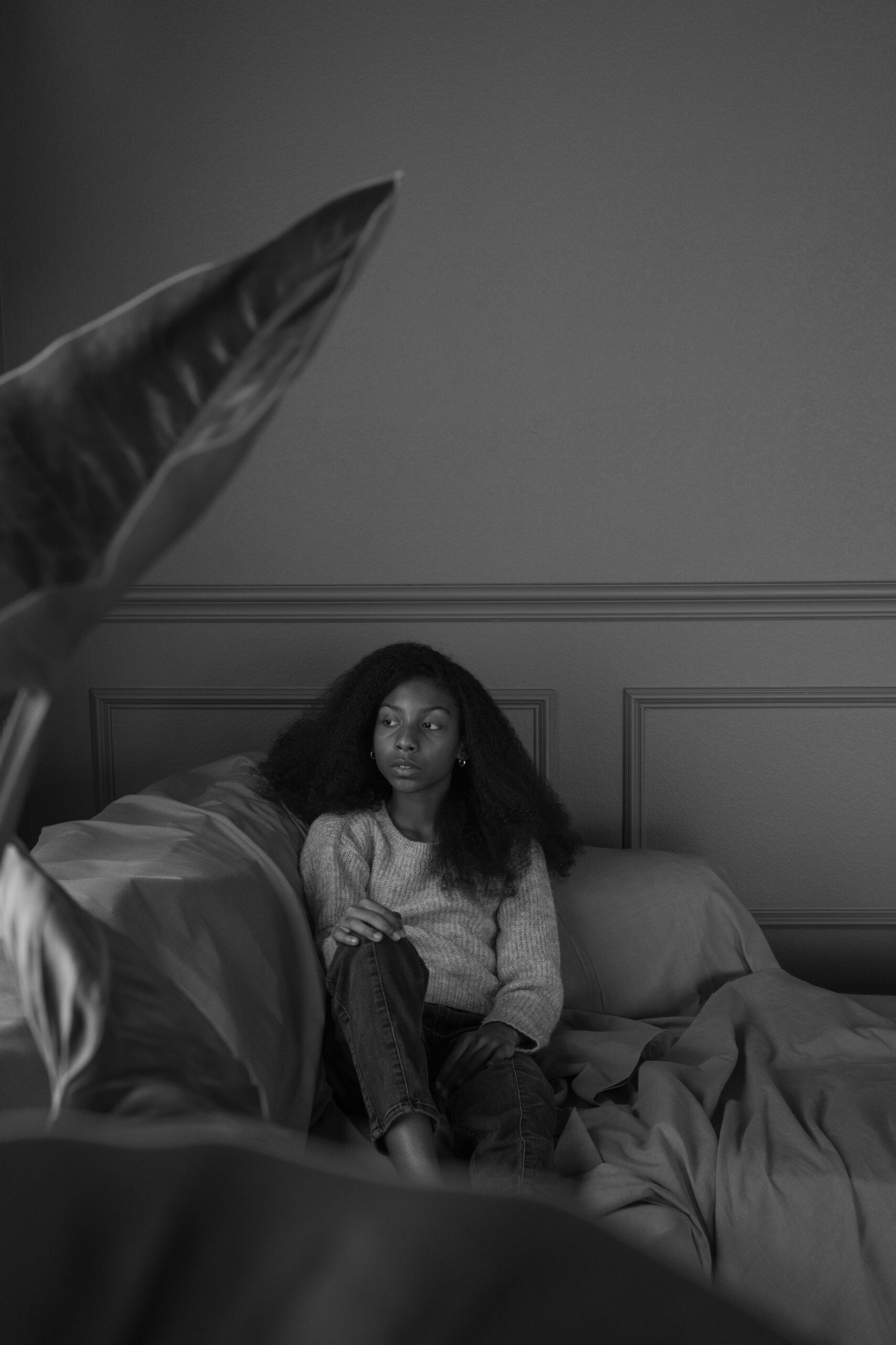 Black-and-white photo of an 11-year-old girl sitting on a couch, surrounded by soft textures and framed by a large houseplant. Her gaze is thoughtful and distant, evoking a sense of calm contemplation and quiet introspection in a serene, intimate setting.