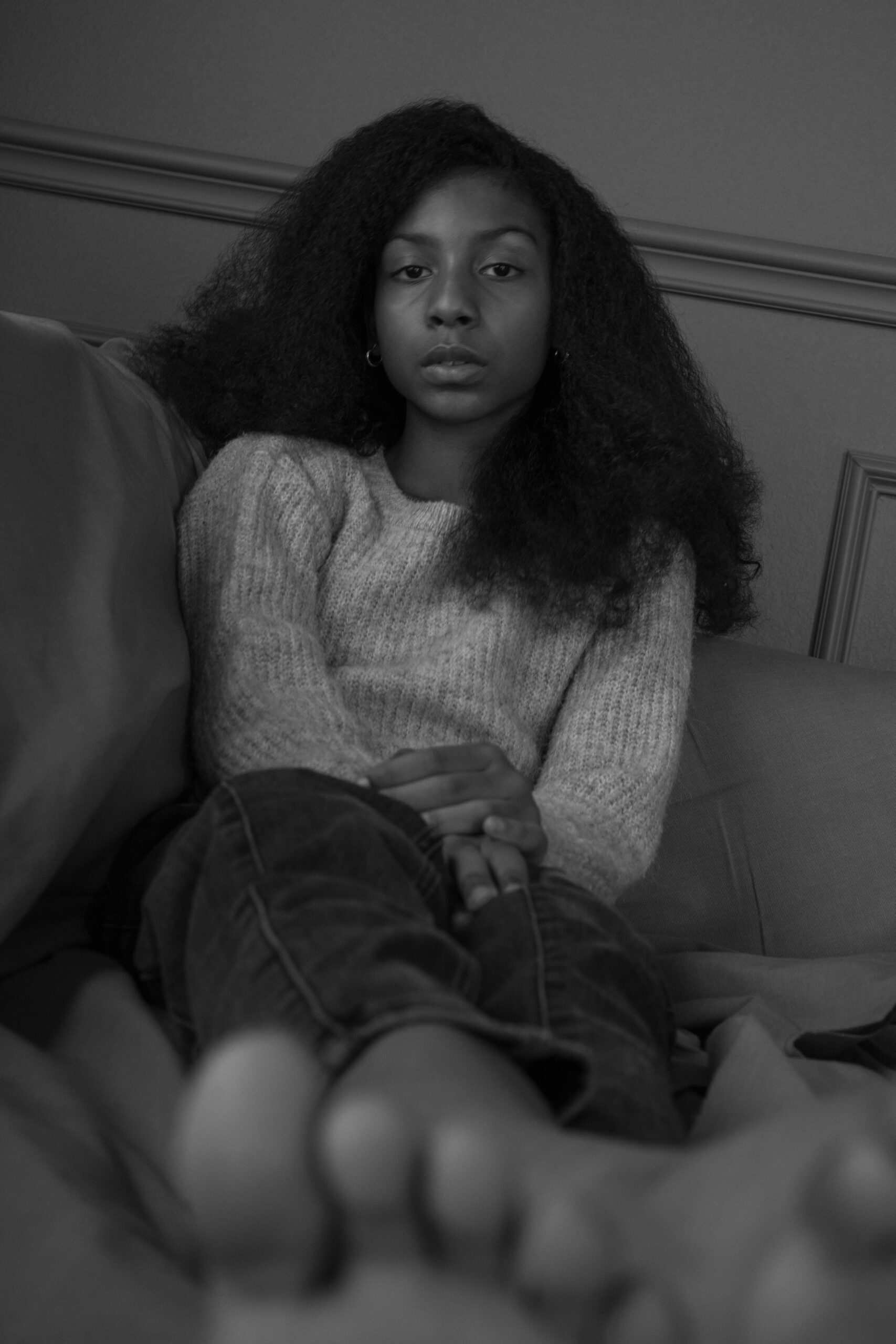 Black-and-white portrait of an 11-year-old girl seated on a couch, her hands gently folded and legs extended. Her calm demeanor and direct gaze evoke a sense of self-assurance and quiet reflection, capturing a moment of serene contemplation.