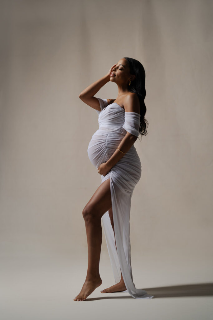 A pregnant woman in a soft, neutral gown posing gracefully in a studio setting, showcasing a classic maternity photoshoot style with timeless and elegant details. A visual reference for how to choose your maternity photoshoot style.