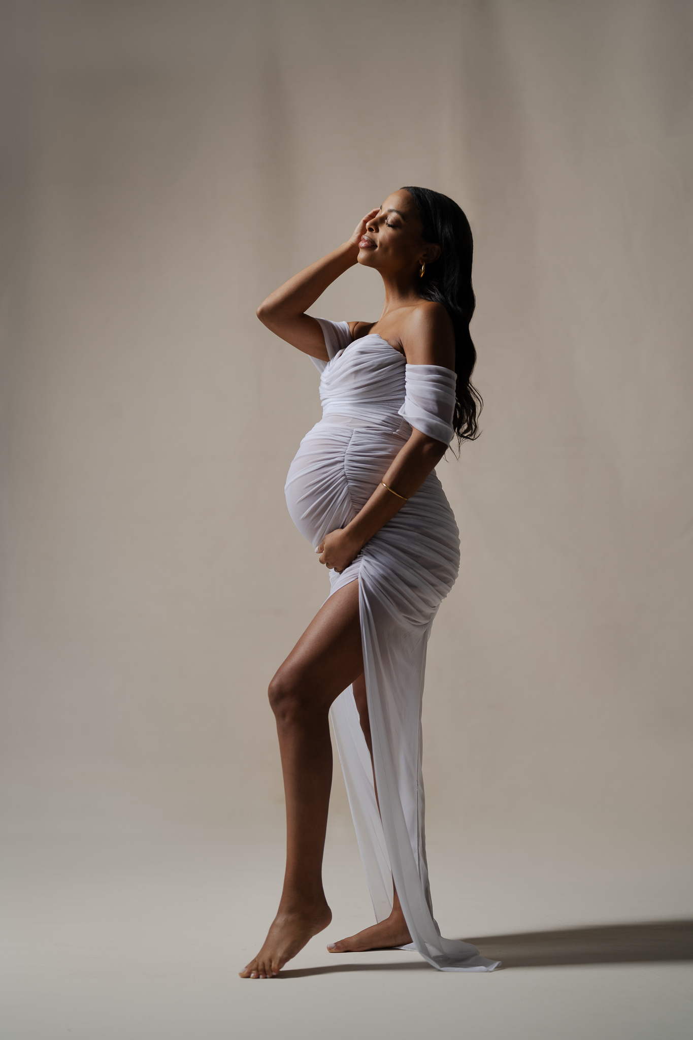 A pregnant woman in a soft, neutral gown posing gracefully in a studio setting, showcasing a classic maternity photoshoot style with timeless and elegant details. A visual reference for how to choose your maternity photoshoot style.