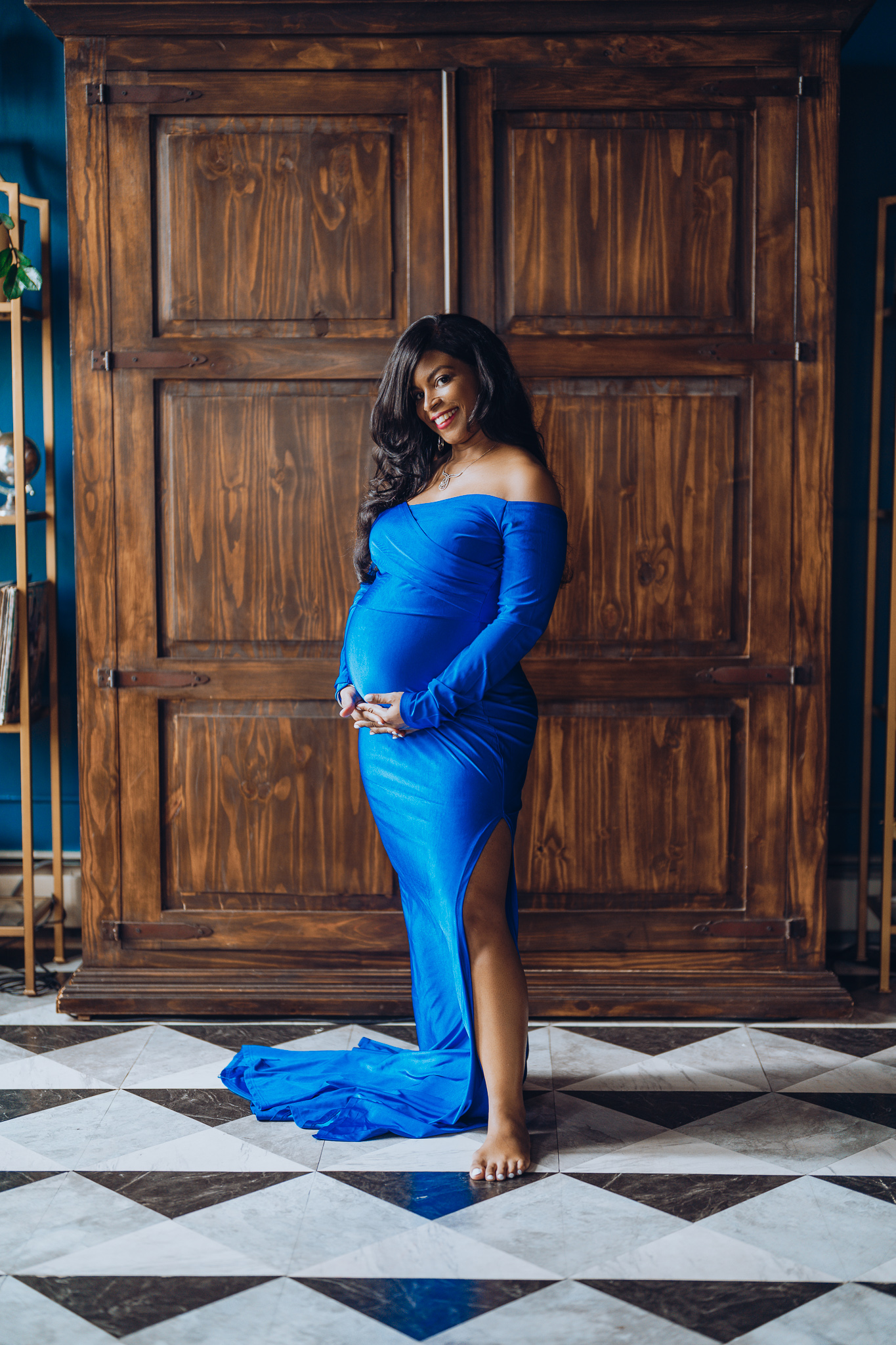Smiling pregnant woman in a royal blue off-the-shoulder gown standing barefoot in a studio with wooden cabinets and patterned tile flooring, showcasing maternity posing tips for a relaxed and beautiful session.